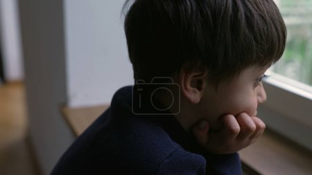 Photo for Thoughtful young boy staring at view from apartment window with hand in chin, pensive child with daydreaming expression - Royalty Free Image