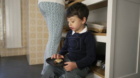 Téléchargez les photos : Petit garçon en train de grignoter du pain avec du fromage assis sur le sol de la cuisine et appuyé sur des meubles tandis que la mère se tient dans la cuisine de fond - en image libre de droit