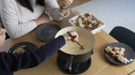Photo for Homemade Swiss Fondue Delight, Mother and child Savoring Cheese and Bread at home mealtime. People eating traditional Swiss dish - Royalty Free Image