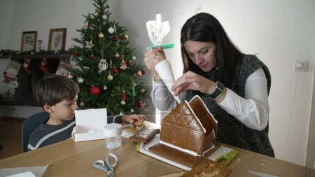 Photo for Mother creating gingerbread house with her son, applying royal ice. Family preparing for december holidays at home - Royalty Free Image