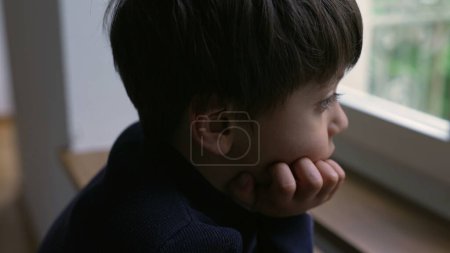 Foto de Niño reflexivo mirando a la vista desde la ventana del apartamento con la mano en la barbilla, niño pensativo con expresión de ensueño - Imagen libre de derechos