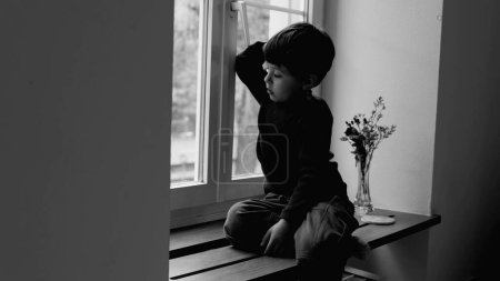 Téléchargez les photos : Petit garçon triste ennuyé assis par la fenêtre regardant dehors dans monochrome exprimant la tristesse et la dépression de l'enfance, noir et blanc - en image libre de droit