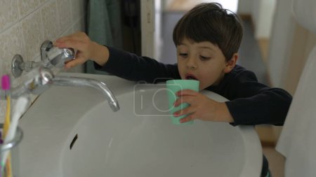 Téléchargez les photos : L'enfant boit du robinet d'eau dans la salle de bain en utilisant une tasse en plastique après s'être brossé les dents pendant le rituel du matin, capturant un style de vie familial quotidien authentique - en image libre de droit