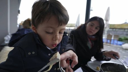 Foto de Niño soplando comida caliente mientras come fondue tradicional suiza en el restaurante con mamá. Padres y niños disfrutando de la comida de la temporada de invierno en el restaurante al aire libre, niños enfriando queso con pan - Imagen libre de derechos