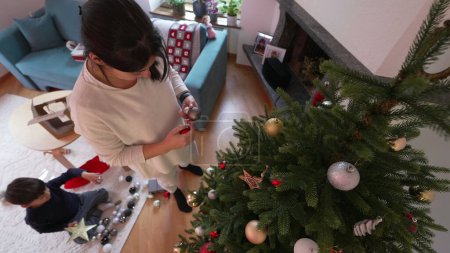 Téléchargez les photos : Mère décorant l'arbre de Noël pendant les vacances d'hiver, femme mettant des boules d'ornement, vue de dessus perspective de la famille se préparant pour les festivités - en image libre de droit