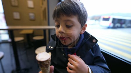 Foto de Niño saboreando el cono de helado en el café sentado junto a la ventana con vistas a la ciudad en movimiento. Niño comiendo dulce postre, cara de cerca - Imagen libre de derechos