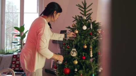 Foto de Mujer cándida decorando el árbol de Navidad durante la temporada navideña en diciembre en casa. Auténtica persona de la vida real añadiendo bolas de adorno, celebrando festividades - Imagen libre de derechos