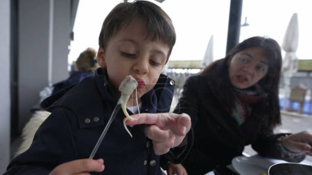 Photo for Child blowing hot food while eating traditional Swiss fondue at restaurant with mom. Parent and little boy enjoying winter season food at outdoor restaurant, kid cooling cheese with bread - Royalty Free Image