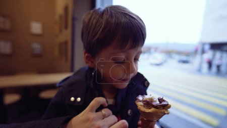 Foto de El niño mancha accidentalmente la bufanda con cono de helado mientras come golosinas en el salón, usando chaqueta. Madre ajusta la ropa del hijo para protegerlo de la mancha - Imagen libre de derechos