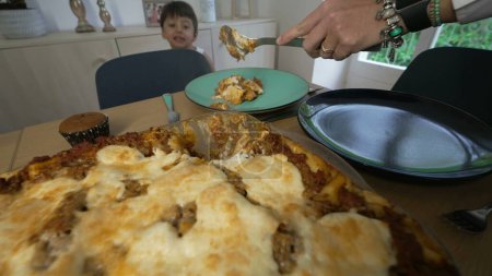 Téléchargez les photos : Gros plan main servant le repas lasagne pour le déjeuner avec l'enfant rebondissant en arrière-plan. Famille se préparant à manger - en image libre de droit