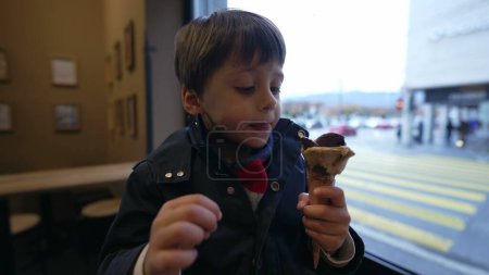 Foto de Niño disfrutando de la delicia de helado sentado junto a la ventana con vistas a la ciudad en el fondo. Niño comiendo dulce postre snack cone - Imagen libre de derechos