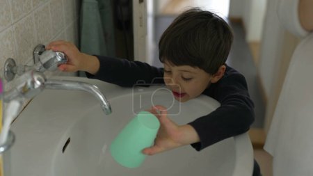 Téléchargez les photos : L'enfant boit du robinet d'eau dans la salle de bain en utilisant une tasse en plastique après s'être brossé les dents pendant le rituel du matin, capturant un style de vie familial quotidien authentique - en image libre de droit