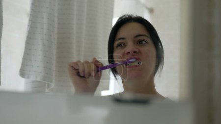 Photo for Woman staring at her own mirror reflection while brushing teeth during morning ritual. Person starting the day routine maintaining dental hygiene during everyday domestic lifestyle - Royalty Free Image