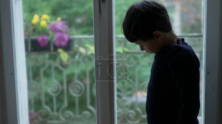 Foto de Niño se para junto a la ventana sintiendo aburrimiento en casa mirando hacia abajo sin nada que hacer - Imagen libre de derechos