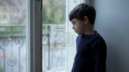 Foto de Niño introspectivo de pie junto a la ventana mira desde el apartamento con triste expresión solitaria y emoción reflexiva - Imagen libre de derechos