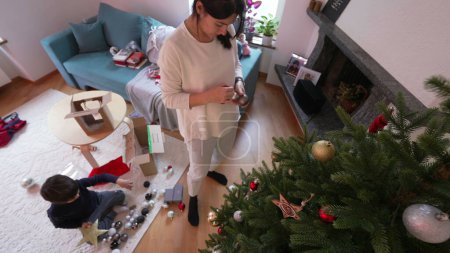 Téléchargez les photos : Mère décorant l'arbre de Noël pendant les vacances d'hiver, femme mettant des boules d'ornement, vue de dessus perspective de la famille se préparant pour les festivités - en image libre de droit