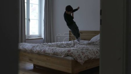 Photo for Child bouncing up and down on top of bed, candid young boy having fun by himself in parent's bedroom by jumping at home feeling energetic - Royalty Free Image