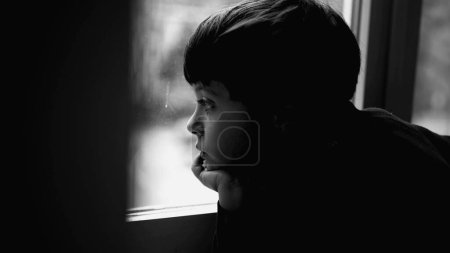 Téléchargez les photos : Enfant déprimé penché sur la fenêtre regardant par la fenêtre tout en luttant contre la maladie mentale, la dépression de l'enfance représentée avec réfléchi jeune garçon en noir et blanc, monochromatique - en image libre de droit
