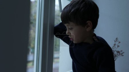 Foto de Niño aburrido atrapado en casa sin nada que hacer, niño pequeño apoyado en la ventana de cristal sintiendo aburrimiento - Imagen libre de derechos