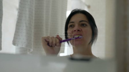 Photo for Woman staring at her own mirror reflection while brushing teeth during morning ritual. Person starting the day routine maintaining dental hygiene during everyday domestic lifestyle - Royalty Free Image