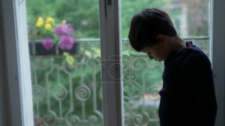 Photo for Child stands by window feeling boredom at home looking down with nothing to do - Royalty Free Image