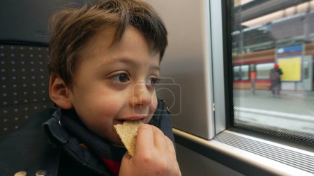 Photo for Young boy snacking cracker while traveling by train staring at platform from European high-speed transportation. Pensive Child eating biscuit while on commute - Royalty Free Image