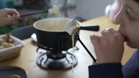 Photo for Child eating fondue, small boy holding fork with piece of bread dipped in Cheese. People enjoying traditional Swiss food at home mealtime - Royalty Free Image