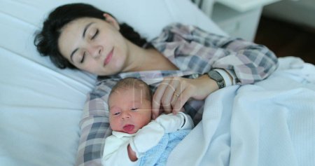 Foto de Mother holding newborn baby falling asleep together at hospital after birth in bed - Imagen libre de derechos