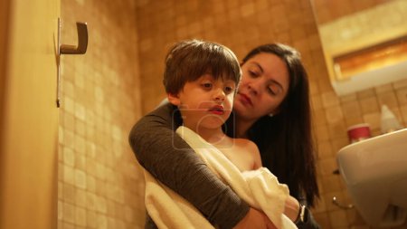 Foto de Madre besar hijo en la mejilla y secado cuerpo envuelto en toalla después de la hora del baño. Padres auténticos y afecto amoroso del niño y relación de cuidado - Imagen libre de derechos