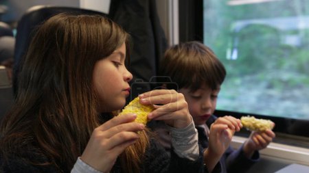 Foto de Children snacking corn inside moving train, siblings - small brother and sister eating healthy food inside high speed transportation - Imagen libre de derechos