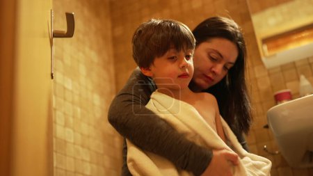 Téléchargez les photos : Mère embrassant son fils sur la joue et séchant le corps enveloppé dans une serviette après l'heure du bain. Authentique parent et enfant affectueux et relation bienveillante - en image libre de droit