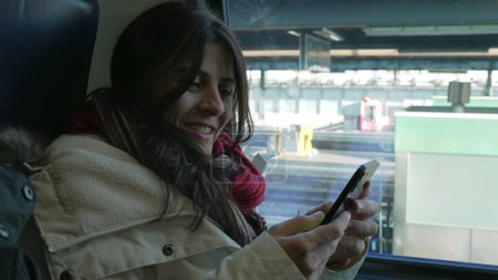 Photo for Commuter Woman Engaged with Smartphone by Train Window. Smiling female passenger holding cellphone device waiting at platform - Royalty Free Image