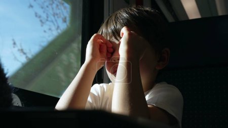 Téléchargez les photos : Exhausted child rubs eyes and face while on a moving train, passenger kid wakes up from nap while traveling feeling tired by window - en image libre de droit