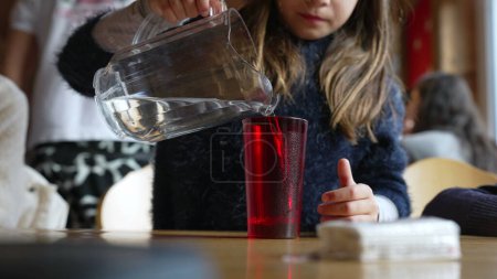 Foto de Niño vertiendo agua en la taza de plástico rojo con el frasco de pie en el restaurante, niña pequeña que sirve agua refrescante del grifo en el comedor - Imagen libre de derechos