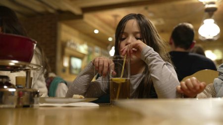 Photo for Little girl drinks ice tea with straw at restaurant in the evening. Child sipping drink at diner - Royalty Free Image