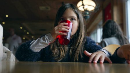 Photo for Small girl seated at restaurant waiting for food to arrive, drinks tap water from red plastic cup and wipes mouth with clothes. Child inside wooden interior - Royalty Free Image