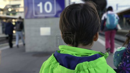 Photo for Back of child walking in train platform station heading to city next to sibling. Young boy walks forward with family wearing raincoat - Royalty Free Image