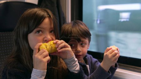 Foto de Niños comiendo comida mientras viajan en tren, niños pasajeros merendando maíz, hermanito y hermana juntos por ventana de transporte - Imagen libre de derechos