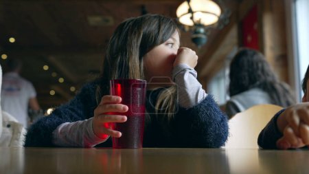 Téléchargez les photos : Petite fille assise au restaurant attendant que la nourriture arrive, boit de l'eau du robinet dans une tasse en plastique rouge et essuie la bouche avec des vêtements. Enfant intérieur en bois intérieur - en image libre de droit