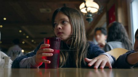 Photo for Small girl seated at restaurant waiting for food to arrive, drinks tap water from red plastic cup and wipes mouth with clothes. Child inside wooden interior - Royalty Free Image