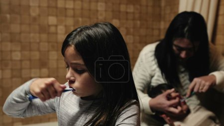 Téléchargez les photos : Routine nocturne avant de se coucher - Les enfants se brossent les dents devant la salle de bain miroir par évier, frères et sœurs avec mère prenant soin de l'hygiène dentaire - en image libre de droit