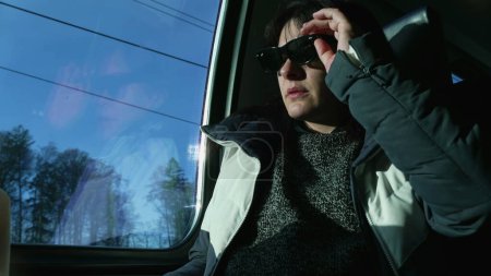 Téléchargez les photos : Woman putting shades while traveling by train, female passenger protecting her eyes by wearing sunglasses inside high speed transportation - en image libre de droit