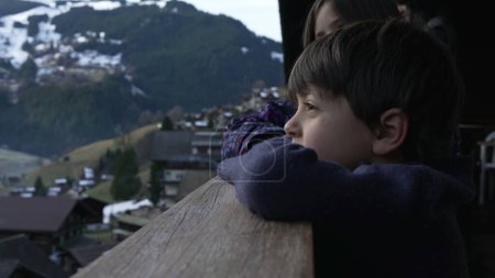 Téléchargez les photos : Enfant penché sur un balcon de chalet alpin en bois regardant le paysage de la montagne pendant la saison d'hiver, vacances en famille dans les Alpes suisses - en image libre de droit