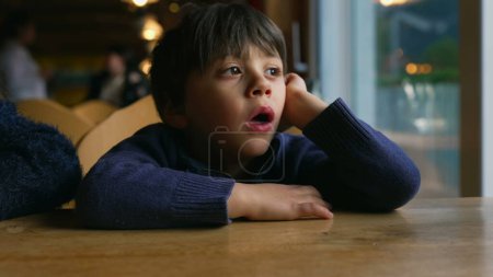 Téléchargez les photos : Petit garçon ennuyé bâillant tout en attendant à la table du restaurant par la fenêtre. Enfant qui se sent ennuyé dans un moment de style de vie authentique en attendant que la nourriture arrive - en image libre de droit