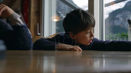 Photo for One small boy at restaurant diner waiting for food to arrive. Child leaning on table by window awaits mealtime - Royalty Free Image