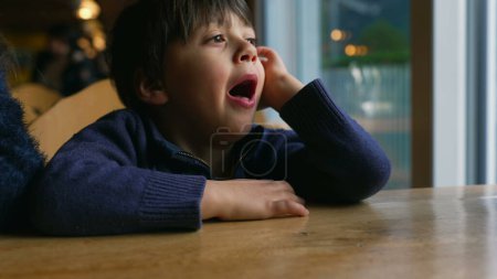 Foto de Niño aburrido bostezando mientras espera en la mesa del restaurante por la ventana. Niño sintiendo aburrimiento en un auténtico momento de estilo de vida esperando a que llegue la comida - Imagen libre de derechos