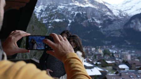 Photo for People vacationing in the Swiss Alps, man taking photo of two women with phone standing in chalet balcony during winter season with mountains in background covered in snow - Royalty Free Image