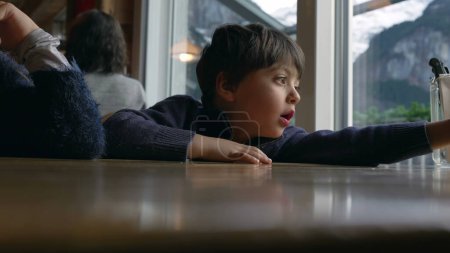 Photo for One small boy at restaurant diner waiting for food to arrive. Child leaning on table by window awaits mealtime - Royalty Free Image