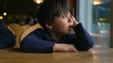 Foto de Niño aburrido bostezando mientras espera en la mesa del restaurante por la ventana. Niño sintiendo aburrimiento en un auténtico momento de estilo de vida esperando a que llegue la comida - Imagen libre de derechos