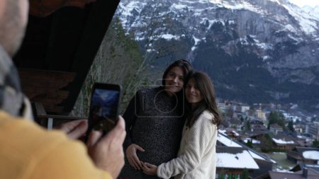 Photo for People vacationing in the Swiss Alps, man taking photo of two women with phone standing in chalet balcony during winter season with mountains in background covered in snow - Royalty Free Image